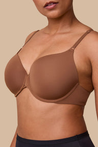 On a neutral beige backdrop, India wears the Base T-Shirt Bra in Brown, shown in a close-up view highlighting the lightly lined cups, signature X-bridge at the center front, and smooth microfiber fabric.