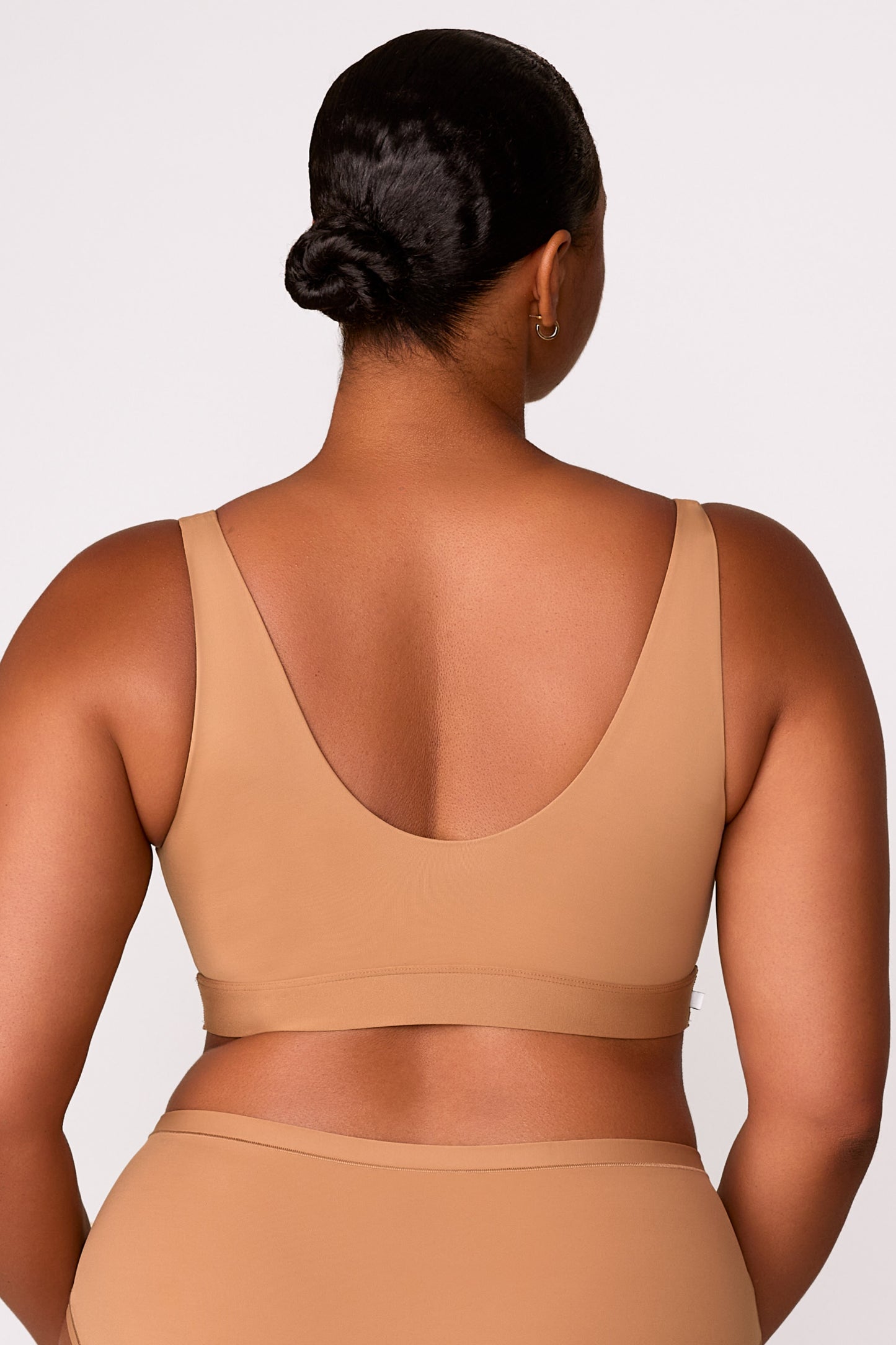 On a white studio backdrop, Zarria wears the Bliss Bralette in Tan, showing the wide straps, and smooth fabric from the back.