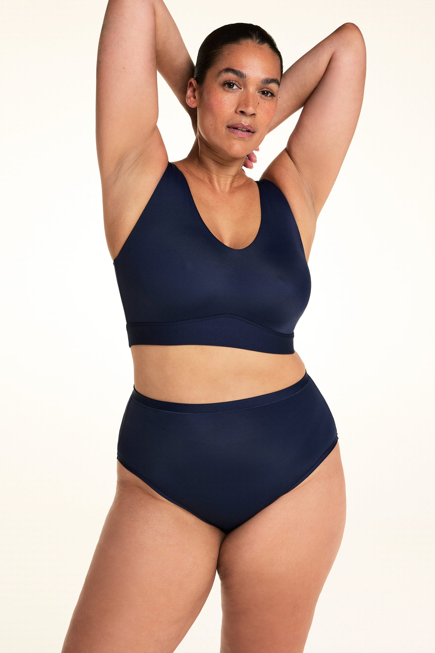 On a light studio backdrop, Jennifer wears the Bliss Highwaist in Navy with a matching navy bralette. Standing with both arms raised, she showcases the high-rise fit, smooth waistband, and comfortable coverage.
