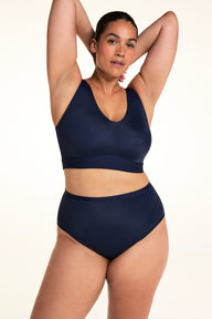 On a light studio backdrop, Jennifer wears the Bliss Highwaist in Navy with a matching navy bralette. Standing with both arms raised, she showcases the high-rise fit, smooth waistband, and comfortable coverage.