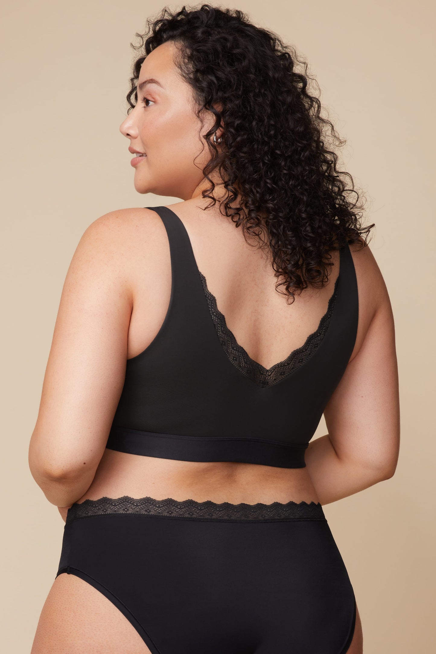 On a neutral beige backdrop, Bianca wears the Bliss Lace Bralette in Black with coordinating underwear, showing the back view with a low scoop back, scalloped lace detail, and smooth supportive band.