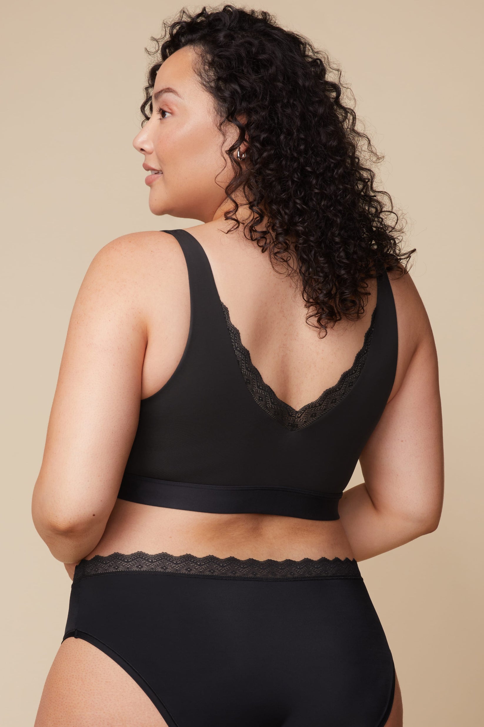 On a neutral beige backdrop, Bianca wears the Bliss Lace Bralette in Black with coordinating underwear, showing the back view with a low scoop back, scalloped lace detail, and smooth supportive band.