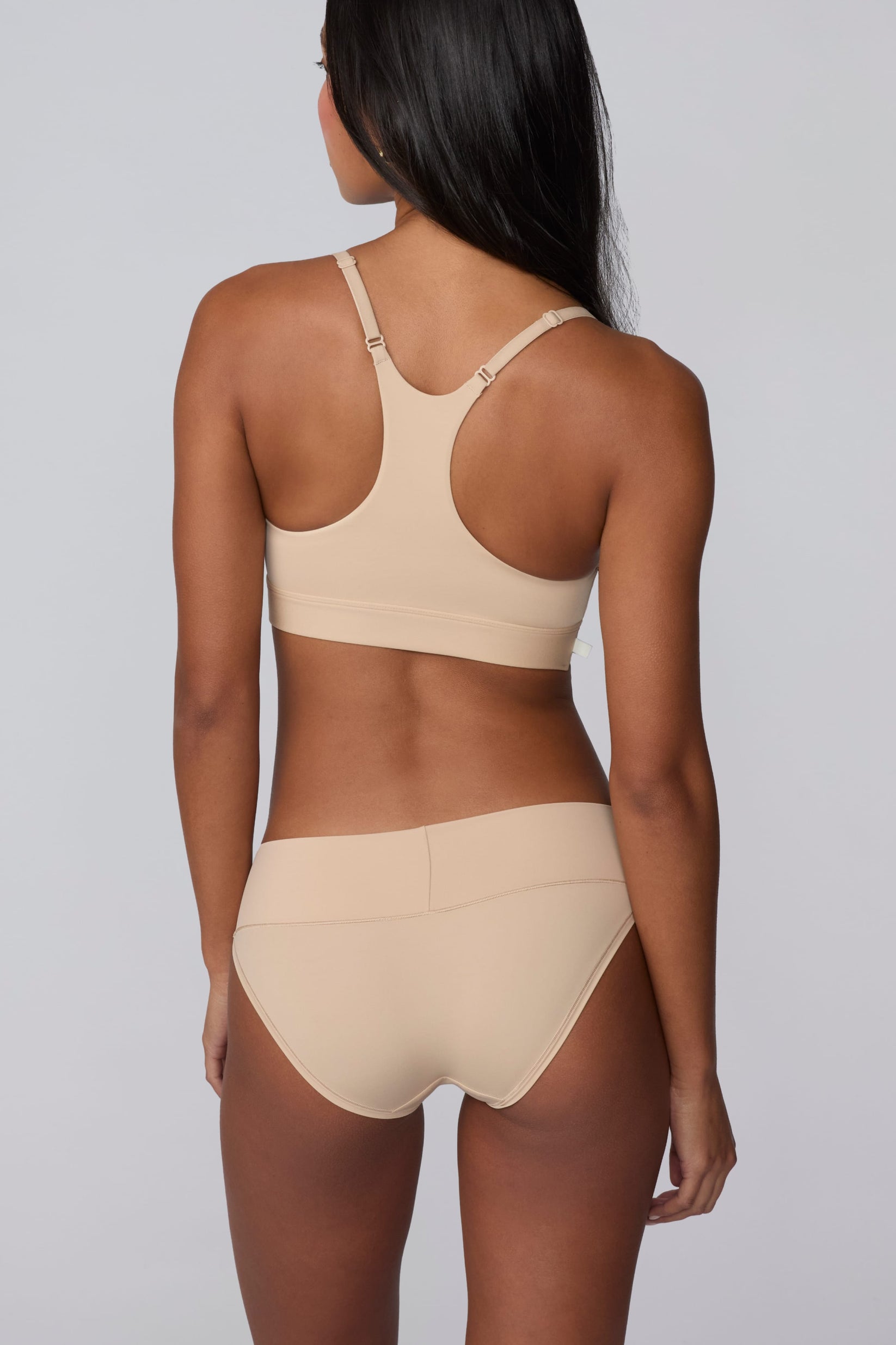In a studio setting, Amber wears the Bliss Low Rise Bikini in Beige with the matching Bliss Bralette in Beige. The back view highlights the racerback design of the bralette and the bikini’s full back coverage.