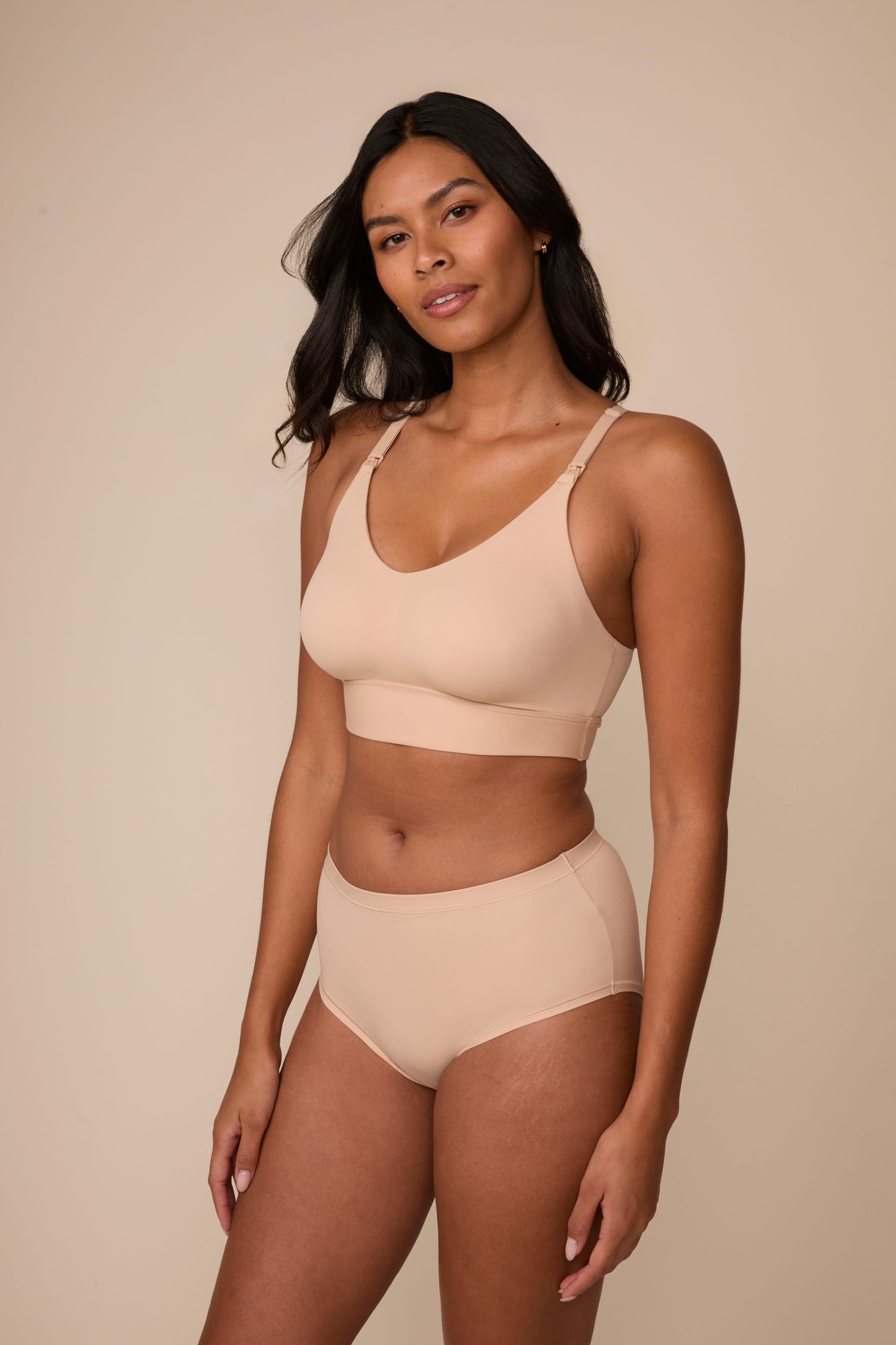 On a neutral studio backdrop, Victoria wears the Bliss Nursing Bralette in Beige with matching underwear. The angled view highlights the scoop neckline, nursing clips, smooth fabric, and supportive underband.