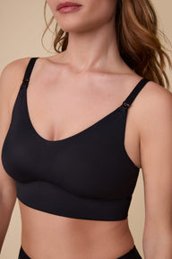 On a neutral beige backdrop, Gracie wears the Bliss Nursing Bralette in Black, highlighting the scoop neckline, nursing clips, smooth fabric, and wireless support.