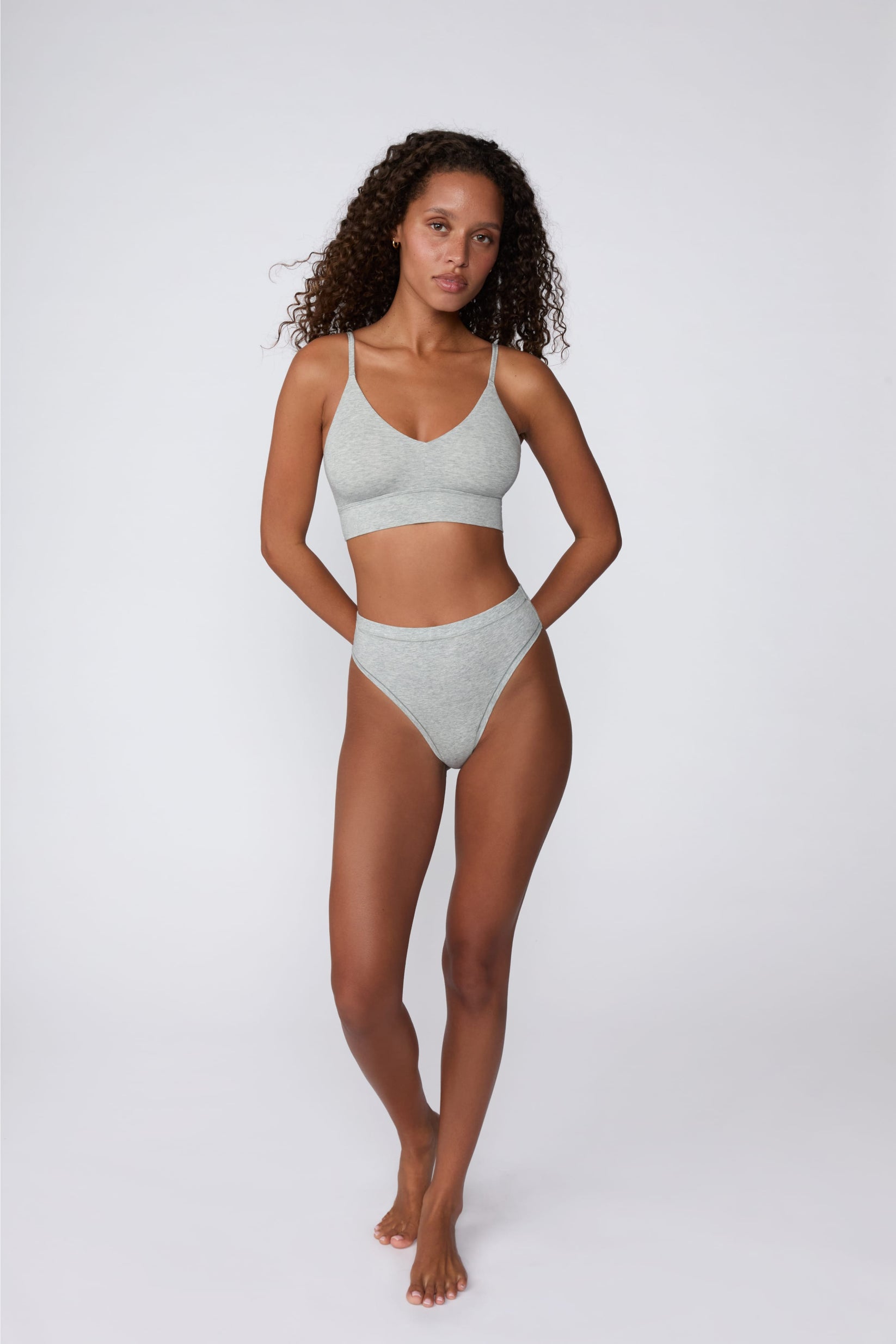 On a light gray studio backdrop, Courtney wears the Cloud Cotton Highwaist Thong in Heather Grey with the matching bralette. The full-body front view highlights the high-rise waistband, high-leg silhouette, and soft cotton fabric.