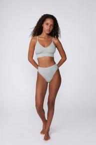 On a light gray studio backdrop, Courtney wears the Cloud Cotton Highwaist Thong in Heather Grey with the matching bralette. The full-body front view highlights the high-rise waistband, high-leg silhouette, and soft cotton fabric.