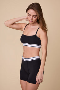 On a neutral beige backdrop, Gracie wears the Cloud Cotton Retro Bandeau Bralette in Black with matching shorts, showcasing the smooth fabric and striped elastic waistband details from an angled view.