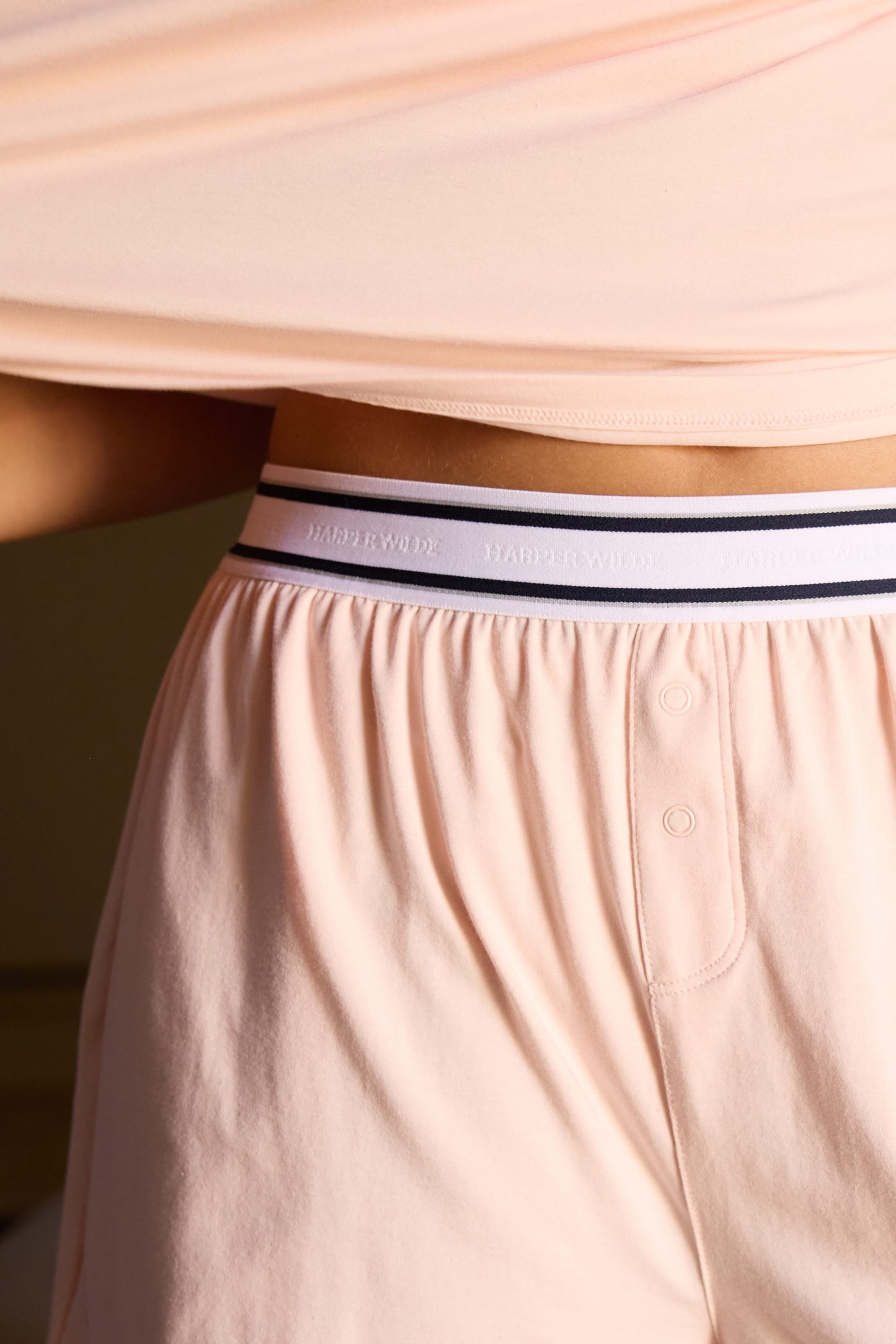 In a softly lit bedroom, Laura wears the Cloud Cotton Short in Dogwood with the Cloud Cotton Tee in Dogwood. The close-up highlights the elastic Harper Wilde logo waistband, functional snap fly detail, and soft fabric.