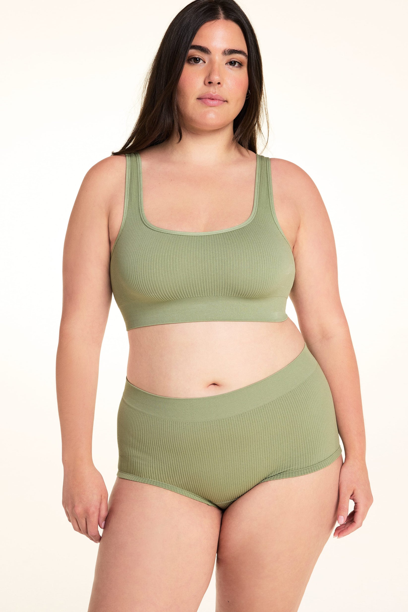 On a white studio backdrop, Alessandra wears the Lounge Rib Scoop Bralette in Dill with the matching Lounge Rib Sleepshort. The front view highlights the scoop neckline, wide straps, ribbed fabric, and supportive underband.