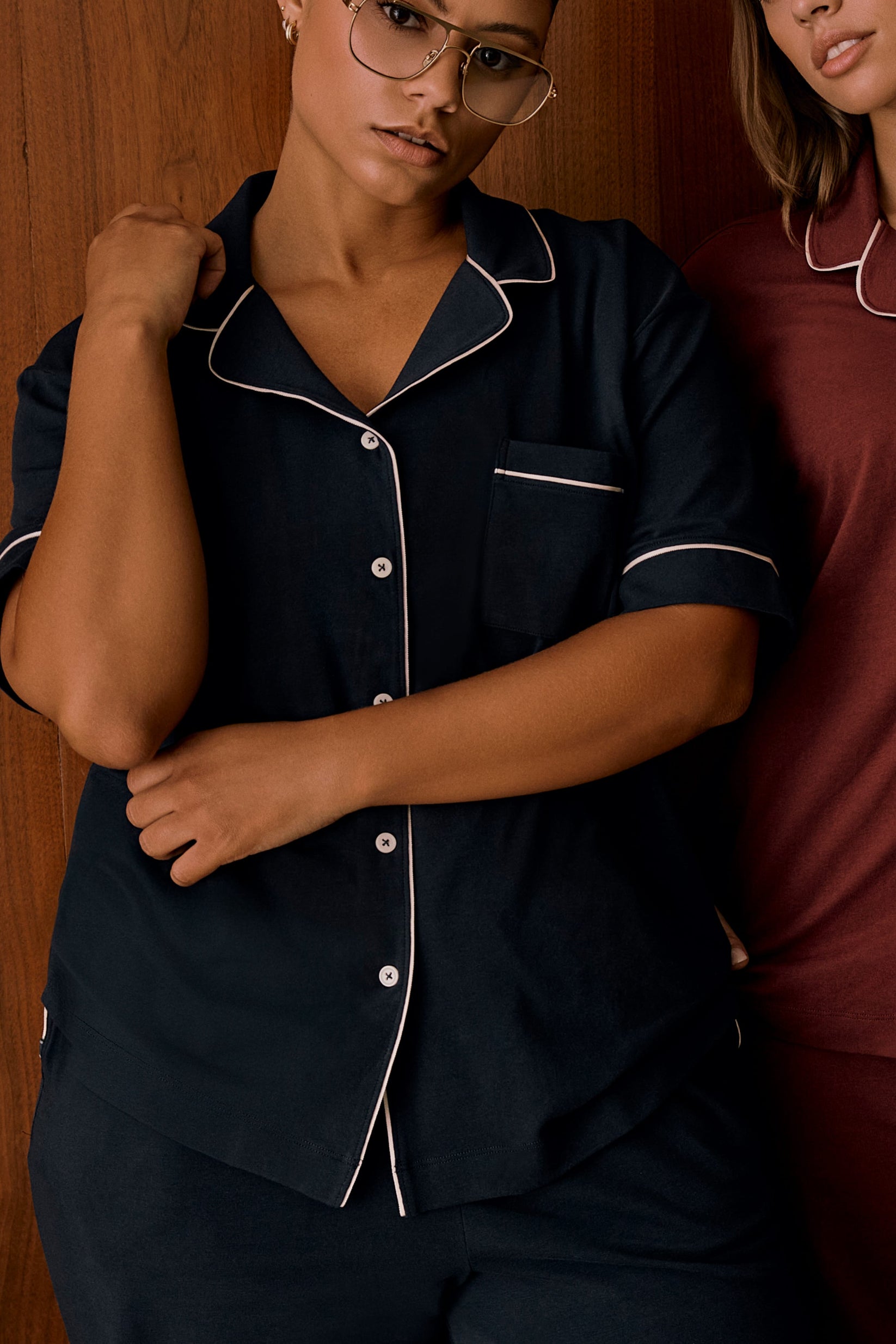 In an indoor setting with a wood-paneled backdrop, India wears the Perfect Pima Pajama Shirt in Navy. The close-up view highlights the button-front closure, chest pocket, contrast piping, and relaxed fit. Another model wearing a burgundy pajama top is partially visible beside her.