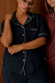 In an indoor setting with a wood-paneled backdrop, India wears the Perfect Pima Pajama Shirt in Navy. The close-up view highlights the button-front closure, chest pocket, contrast piping, and relaxed fit. Another model wearing a burgundy pajama top is partially visible beside her.