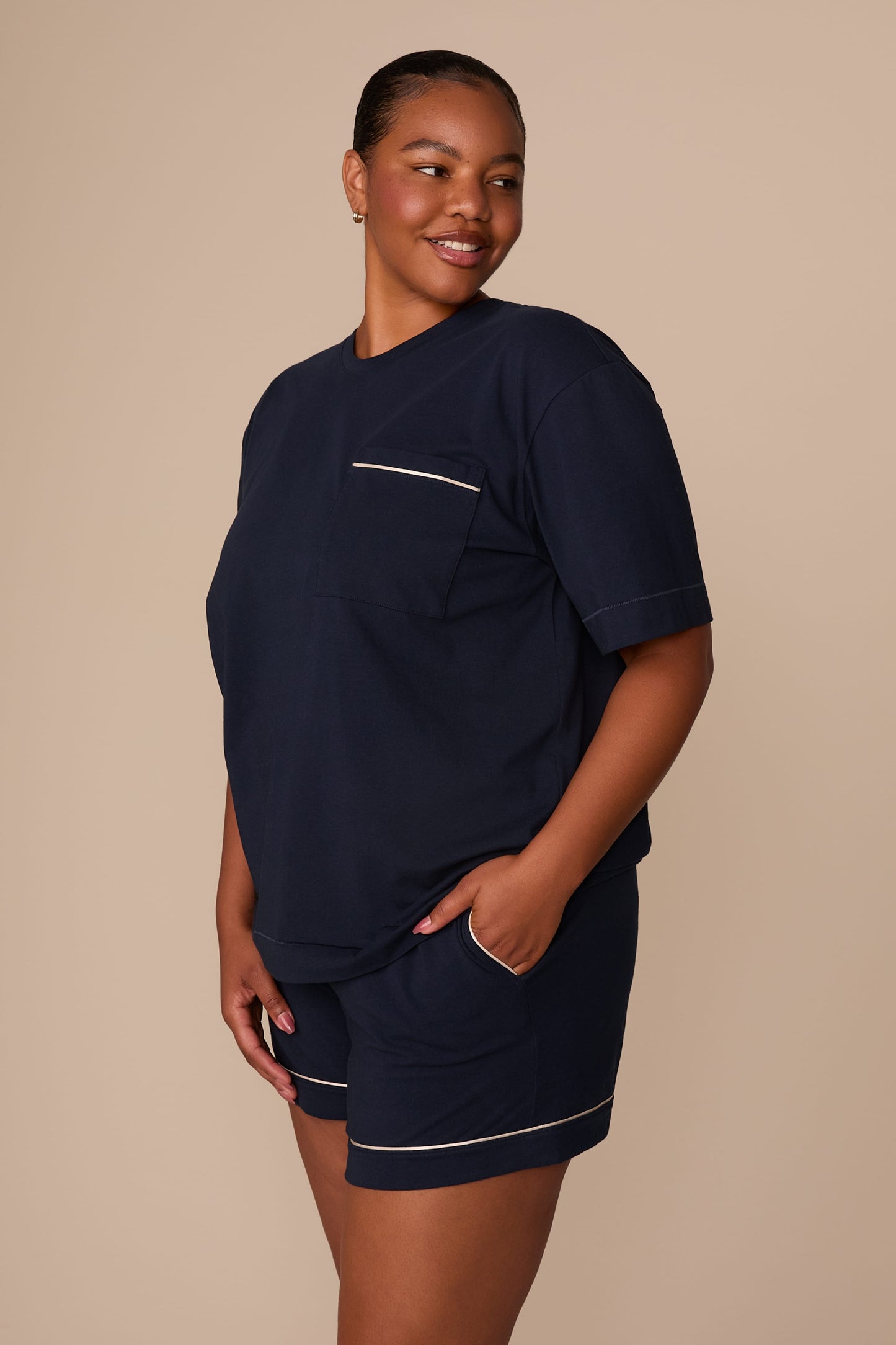 On a warm beige studio backdrop, Zarria wears the Perfect Pima Pajama T-Shirt in a deep navy color with matching pajama shorts. The side-angle view showcases the relaxed silhouette, short sleeves, and chest pocket with contrast piping detail.