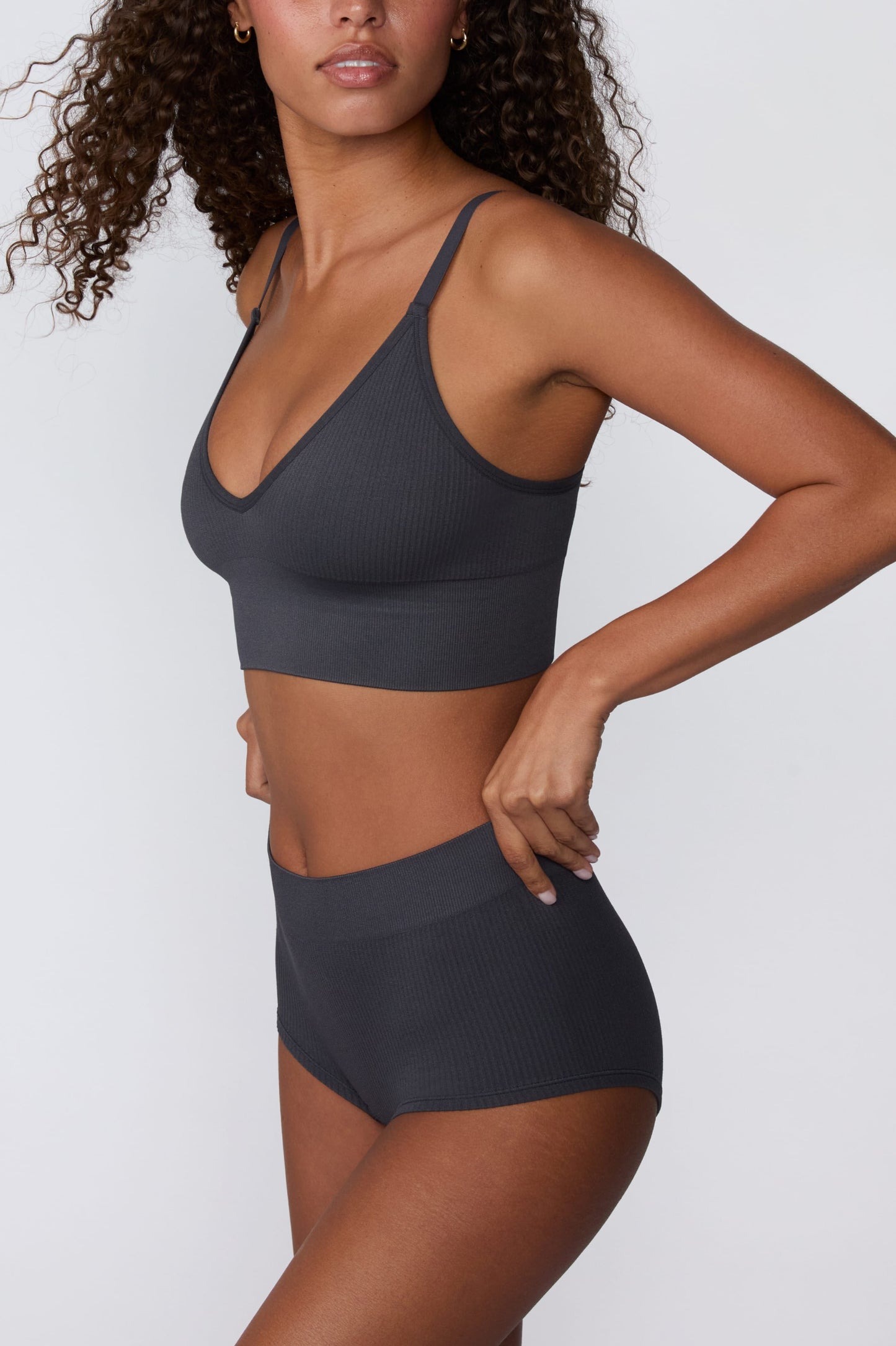 In a studio setting, Courtney wears the Ribbed Seamless Boyshort in Charcoal with a matching ribbed bralette. A side view highlights the high-rise waistband, full-coverage boyshort silhouette, and ribbed texture.
