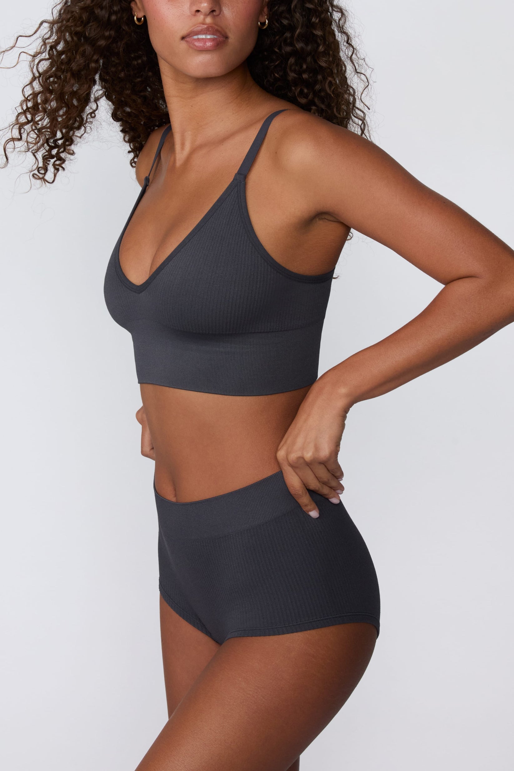 In a studio setting, Courtney wears the Ribbed Seamless Boyshort in Charcoal with a matching ribbed bralette. A side view highlights the high-rise waistband, full-coverage boyshort silhouette, and ribbed texture.