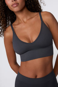 On a light gray studio backdrop, Courtney wears the Ribbed Seamless Triangle Bralette in Charcoal with a matching ribbed bottom. A close-up angled view highlights the ribbed texture, V-neckline, adjustable straps, and wide underband.