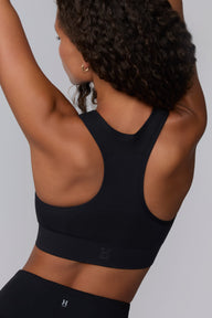 On a light gray studio backdrop, Courtney wears the Seamless Racerback Sports Bra in Black with matching bike shorts. The close-up back view highlights the racerback silhouette, breathable knit texture, wide underband, and tonal Harper Wilde logo at the center back.