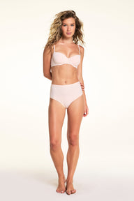 On a light neutral backdrop, Alexis wears the Base Balconette Bra in Rose with matching high-rise underwear. Standing in a full-body pose, the image highlights the balconette neckline, molded underwire cups, adjustable straps, and smooth silhouette.
