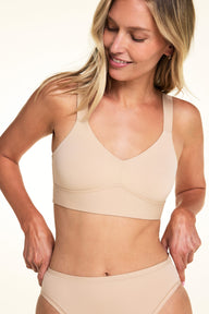On a light neutral backdrop, Meagan wears the Bliss Smoothing Bralette in Beige with matching underwear. She looks down while adjusting the waistband, highlighting the V-neckline, wide straps, and smooth microfiber fabric.