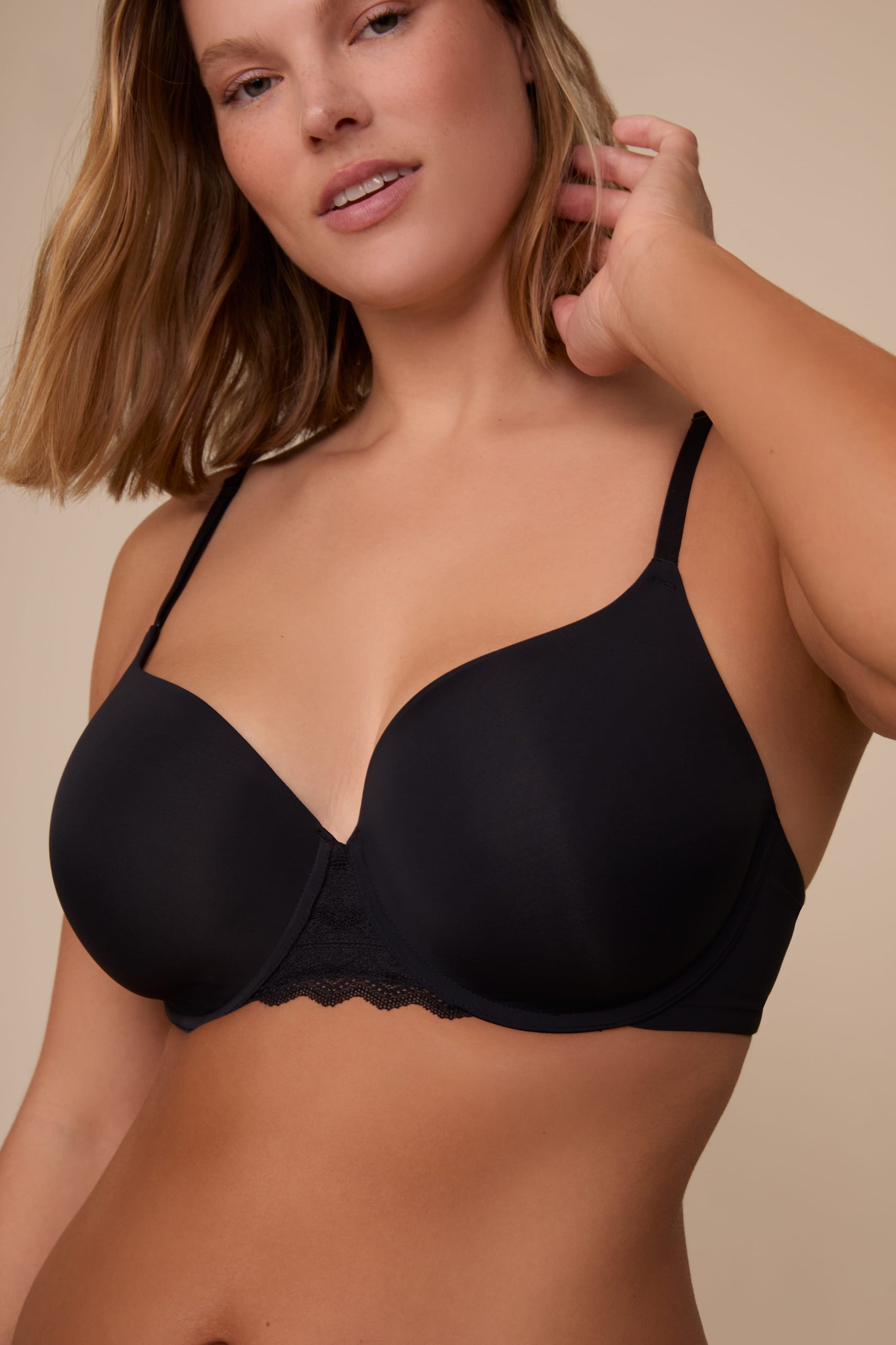 On a neutral studio backdrop, Sonnie wears the Base Lace T-Shirt Bra in Black. The close-up front view highlights the smooth molded cups, underwire support, adjustable straps, and lace detail at the center front.
