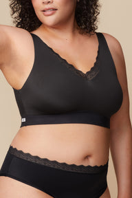 On a neutral beige backdrop, a close-up of Bianca wearing the Bliss Lace Bralette in Black highlights the deep V-neckline, delicate lace trim, smooth fabric, and supportive underband.