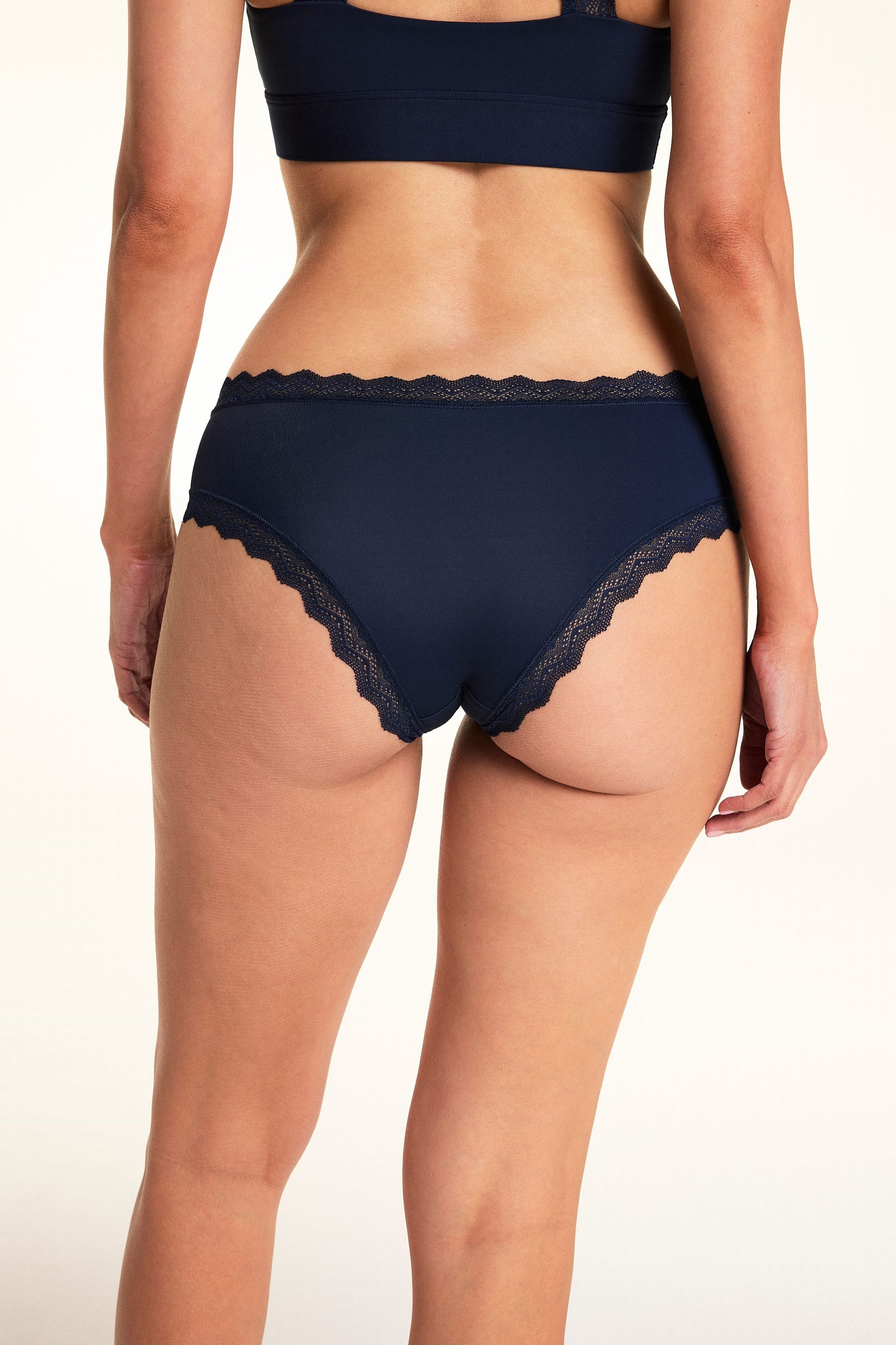 On a light studio backdrop, Meagan is shown from the back wearing the Bliss Lace Cheeky in Navy with the Bliss Lace Halterneck Bralette in Navy. The angled view showcases the cheeky back coverage and lace trim details.