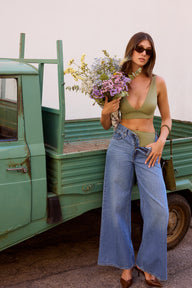 Outdoors beside a vintage green pickup truck, Laura wears the Bliss Plunge Adjustable Bralette in Dill with light-wash jeans worn low on the hips, sunglasses, and carries a bouquet of flowers. The bralette features a deep V-neck, adjustable straps, and a smooth underband for comfortable support.
