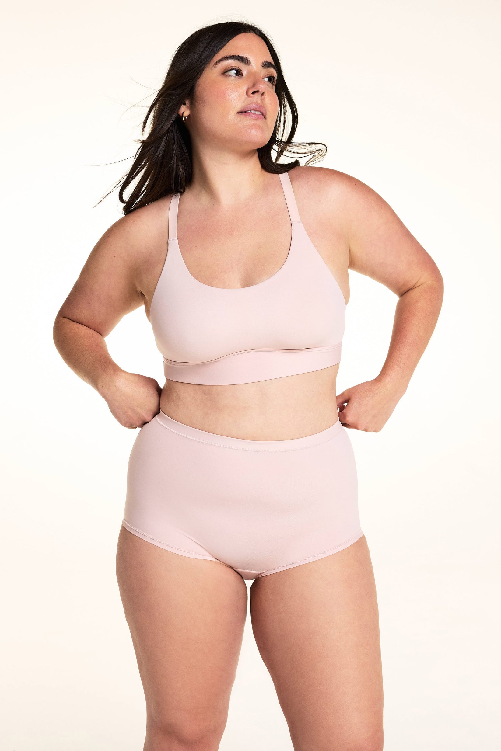 On a light studio backdrop, Alessandra wears the Bliss Racerback Bralette in Ember Rose with matching high-rise bottoms. The full-body image highlights the scoop neckline, smooth fabric, adjustable straps, and wide comfort band.