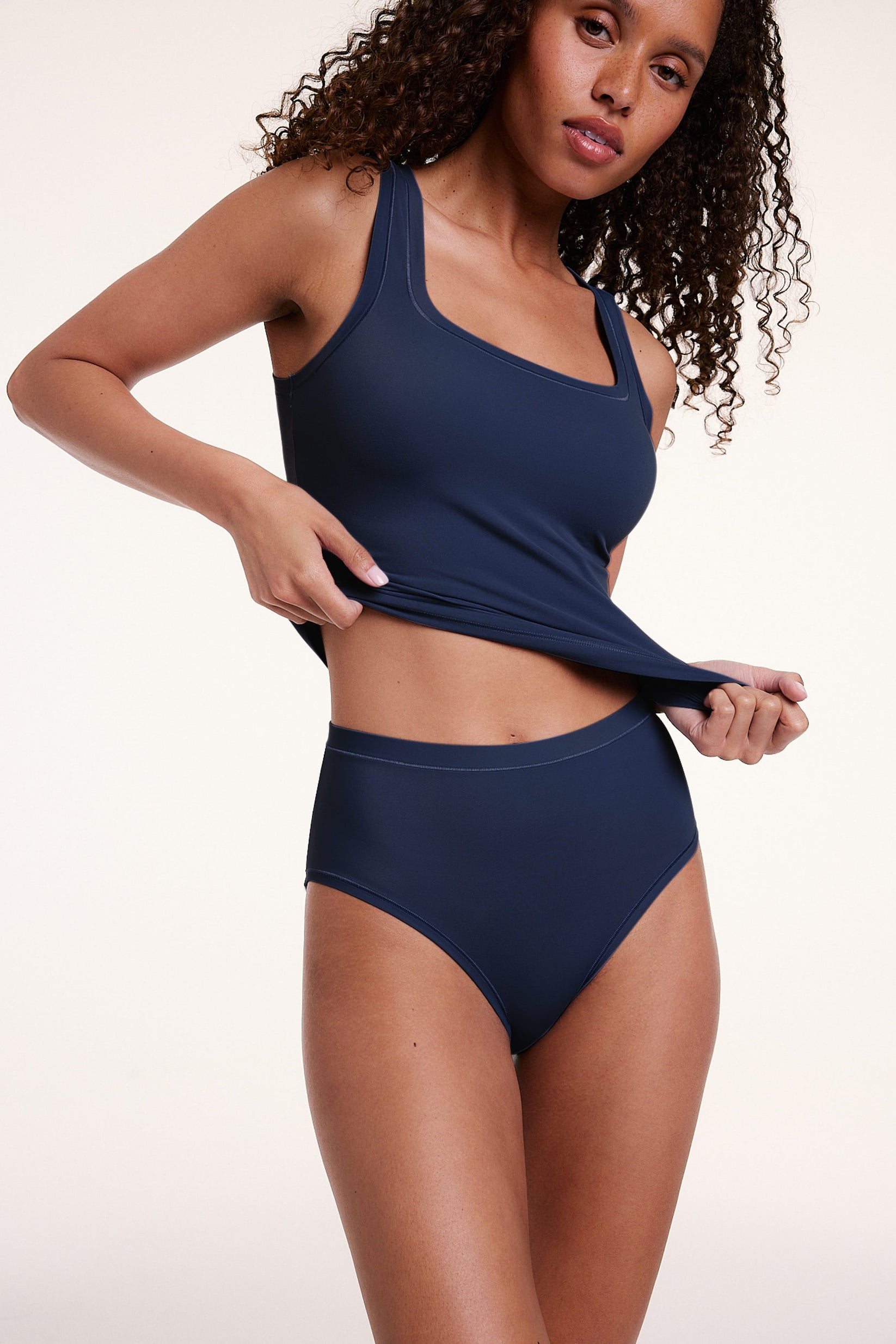 On a light studio backdrop, Courtney wears the Bliss Tank in Navy with matching Navy underwear. She stands with her hands at her hips, showcasing the square neckline, wide straps, and smooth fabric.