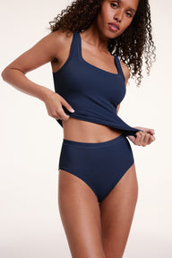 On a light studio backdrop, Courtney wears the Bliss Tank in Navy with matching Navy underwear. She stands with her hands at her hips, showcasing the square neckline, wide straps, and smooth fabric.