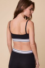 On a neutral beige backdrop, Gracie wears the Cloud Cotton Retro Bandeau Bralette in Black with matching shorts, highlighting the back view, adjustable straps, and striped elastic underband.