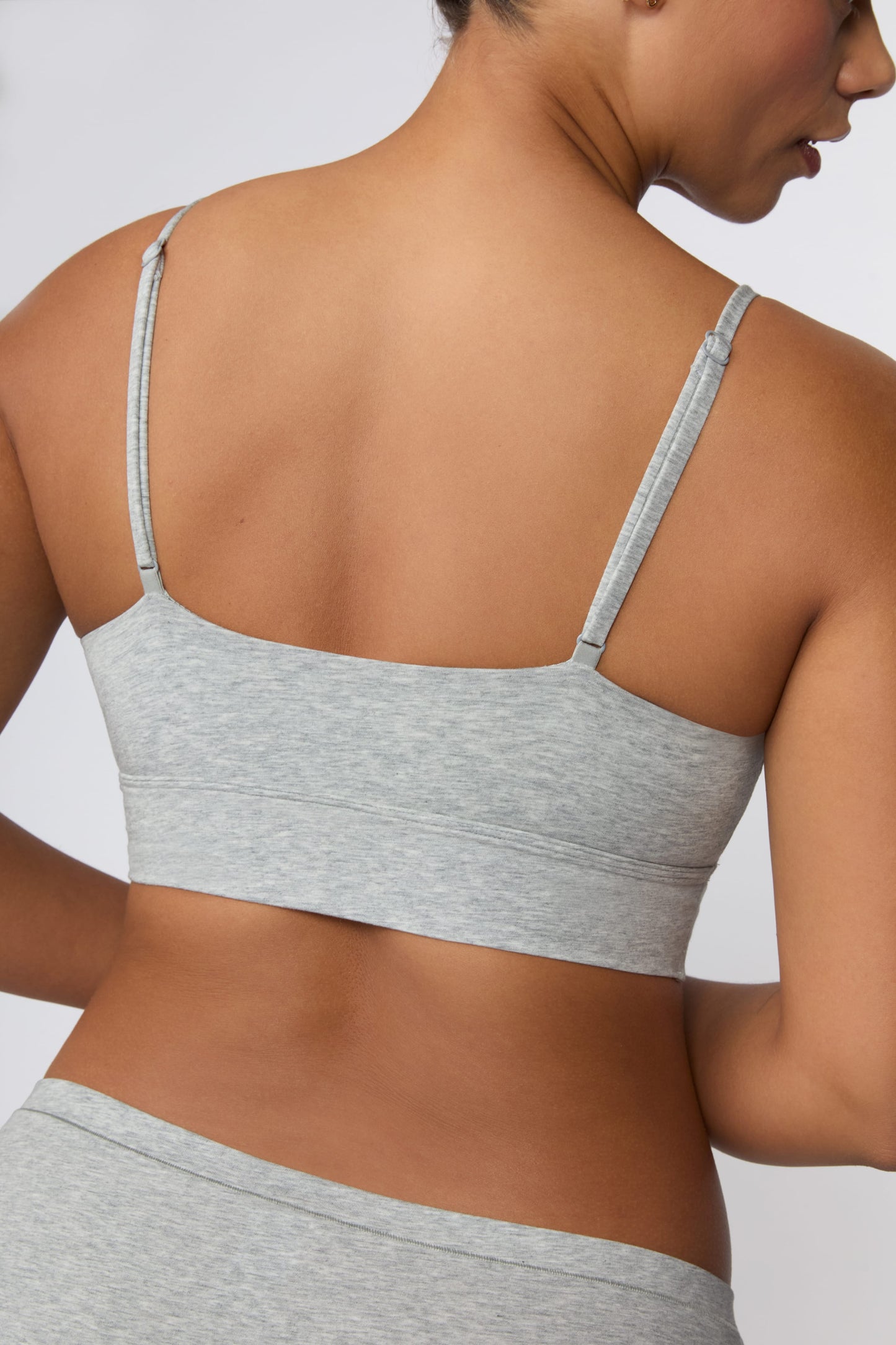 On a light gray studio backdrop, India wears the Cloud Cotton Thin Strap Bralette in Heather Grey with matching underwear. The angled back view highlights the adjustable straps, straight-back silhouette, and wide underband.