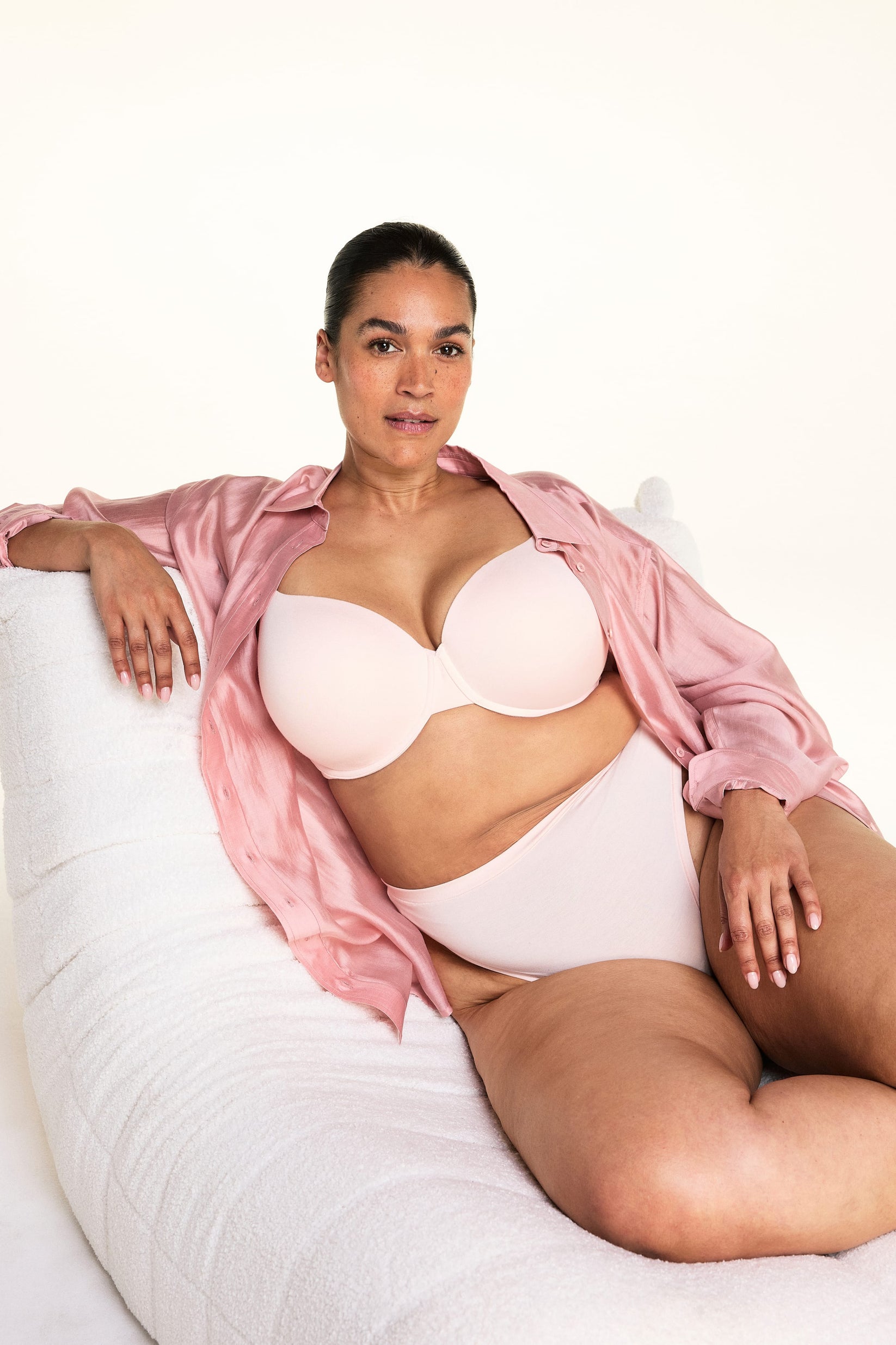 On a light studio backdrop, Jennifer wears the Cloud Cotton Highwaist Thong in Dogwood with a matching bra in Dogwood while seated in a plush white chair and wearing an open pink button-down shirt. The image highlights the high-rise waistband and soft cotton fabric.