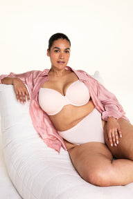 On a light studio backdrop, Jennifer wears the Cloud Cotton Highwaist Thong in Dogwood with a matching bra in Dogwood while seated in a plush white chair and wearing an open pink button-down shirt. The image highlights the high-rise waistband and soft cotton fabric.
