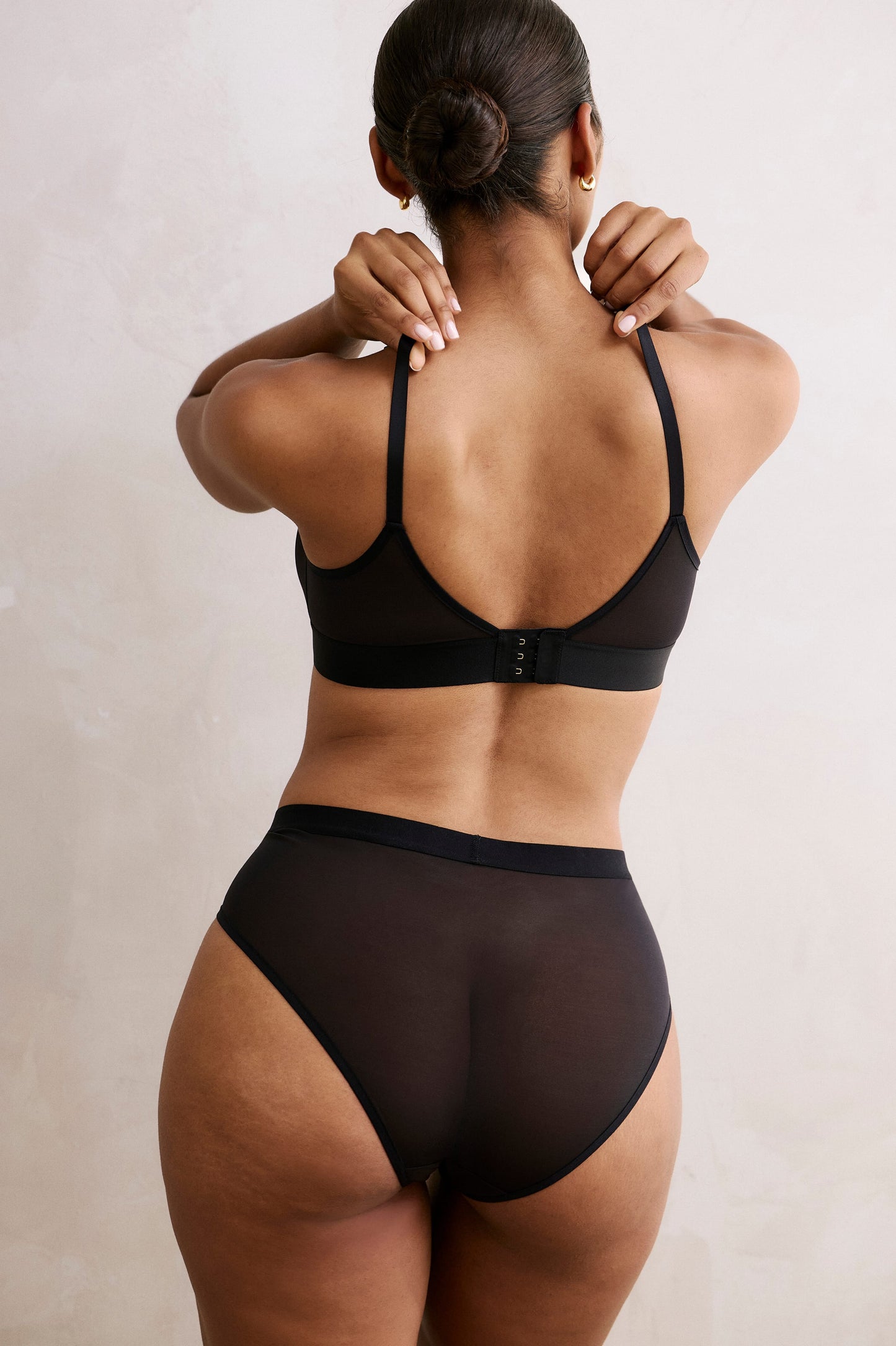 In a softly lit room, back view of a model wearing the ComfortMesh™ Highwaist Brief in Black with the matching ComfortMesh™ Triangle Bralette, showing the sheer mesh fabric and moderate back coverage.