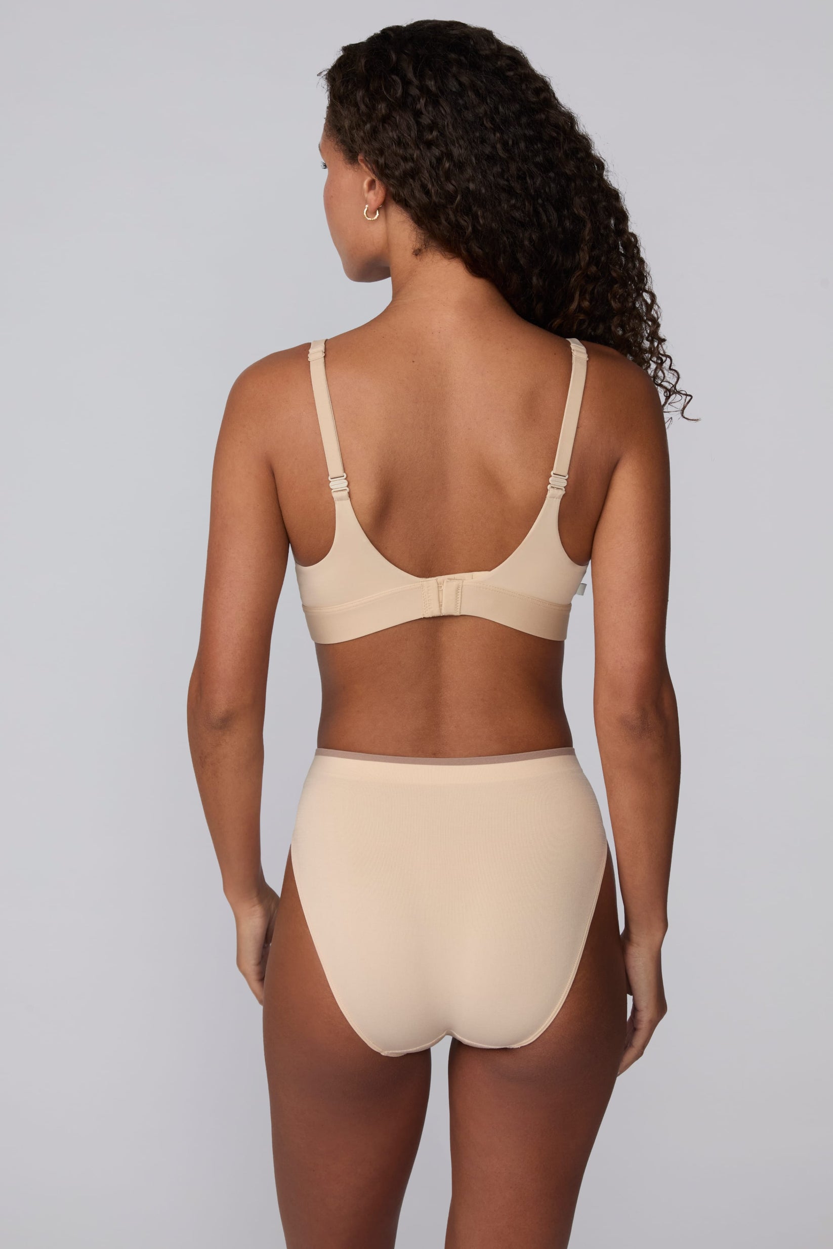 On a light gray studio backdrop, Courtney wears the Smooth Seamless Brief in Beige with the matching Smooth Seamless Bralette in Beige. The back view highlights the full back coverage, smooth finish, and contrast trim waistband.