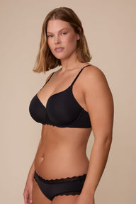 On a neutral studio backdrop, Sonnie wears the Base Lace T-Shirt Bra in Black with matching underwear. The side-front view showcases the smooth molded cups, underwire support, adjustable straps, and lace trim details on the bra and underwear.