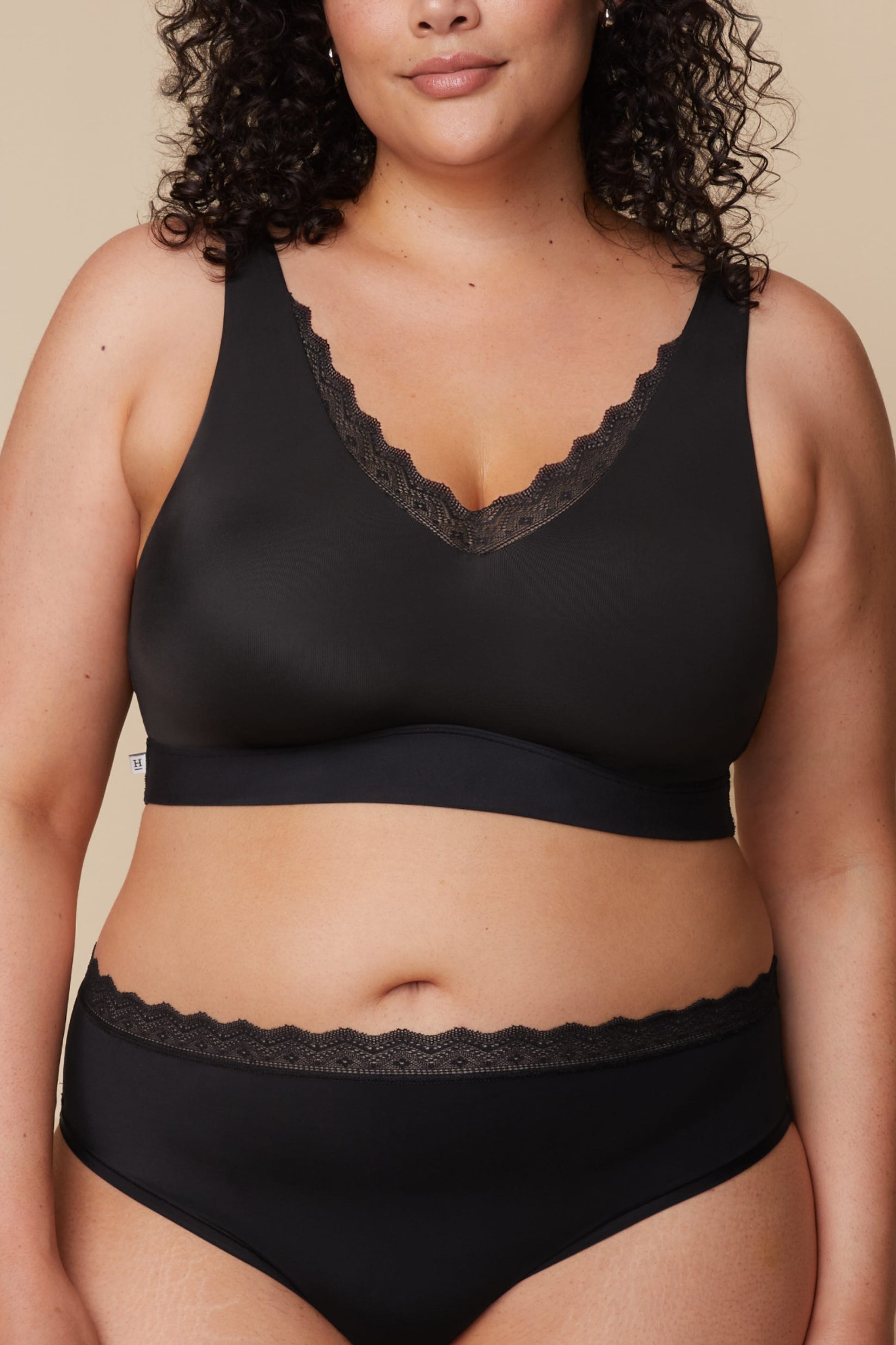 On a neutral beige backdrop, a front-facing close-up of Bianca wearing the Bliss Lace Bralette in Black with coordinating underwear showcases the deep V-neckline with scalloped lace trim, smooth fabric, and supportive underband.