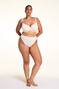 On a light studio backdrop, Jennifer wears the Cloud Cotton Highwaist Thong in Dogwood with a matching bra in Dogwood. The full-body image highlights the high-rise waistband, high-cut leg opening, and soft cotton fabric.