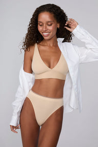 In a studio setting, Courtney wears the Smooth Seamless Brief in Beige with the matching Smooth Seamless Bralette in Beige. An open white button-down shirt drapes off her shoulders while the front view highlights the high-leg silhouette, smooth fabric, and contrast trim waistband.