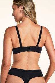 From the back against a white studio backdrop, Meagan wears the Base Strapless Bra in Black with detachable straps and matching thong underwear, showing the hook-and-eye closure and adjustable straps.