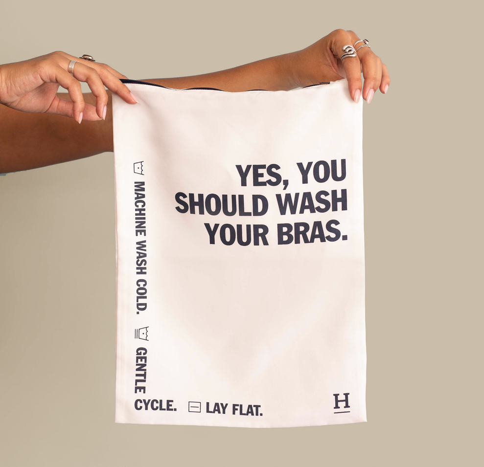 A hand holding a white bra wash bag featuring bold black text that reads, "Yes, you should wash your bras." The bag also displays washing instructions: "Machine wash cold," "Gentle cycle," and "Lay flat."