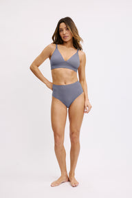 On a white studio backdrop, a model stands wearing the Bliss Triangle Bralette in Steel with the matching high-waist underwear, showing the pullover bralette’s wireless band and soft, supportive fit.