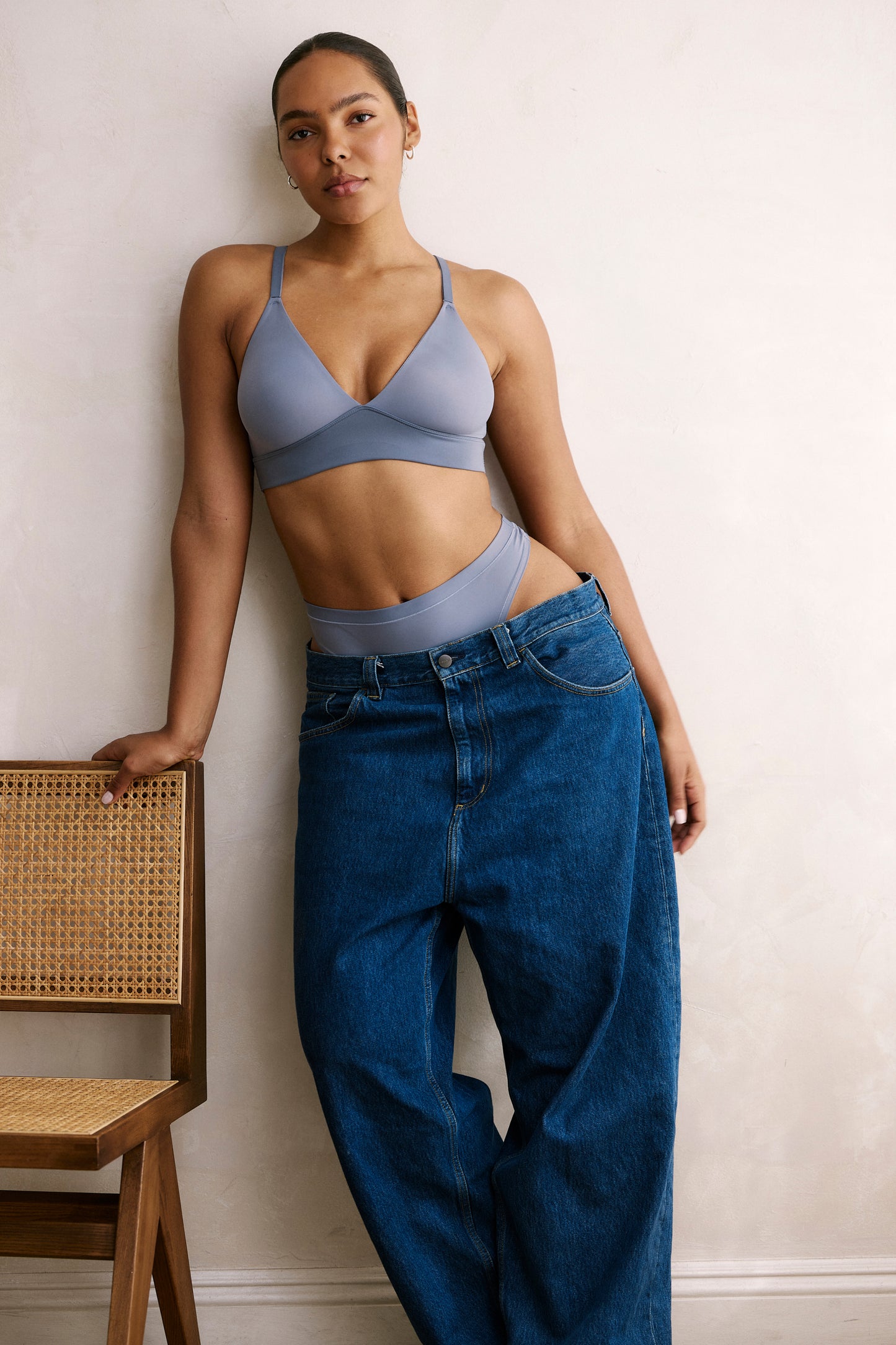 Against a clean white wall, a model wears the Bliss Triangle Bralette in Steel, paired with matching underwear and relaxed denim. The bralette features a deep V neckline, adjustable front straps, and a smooth microfiber band designed for soft, wireless comfort.
