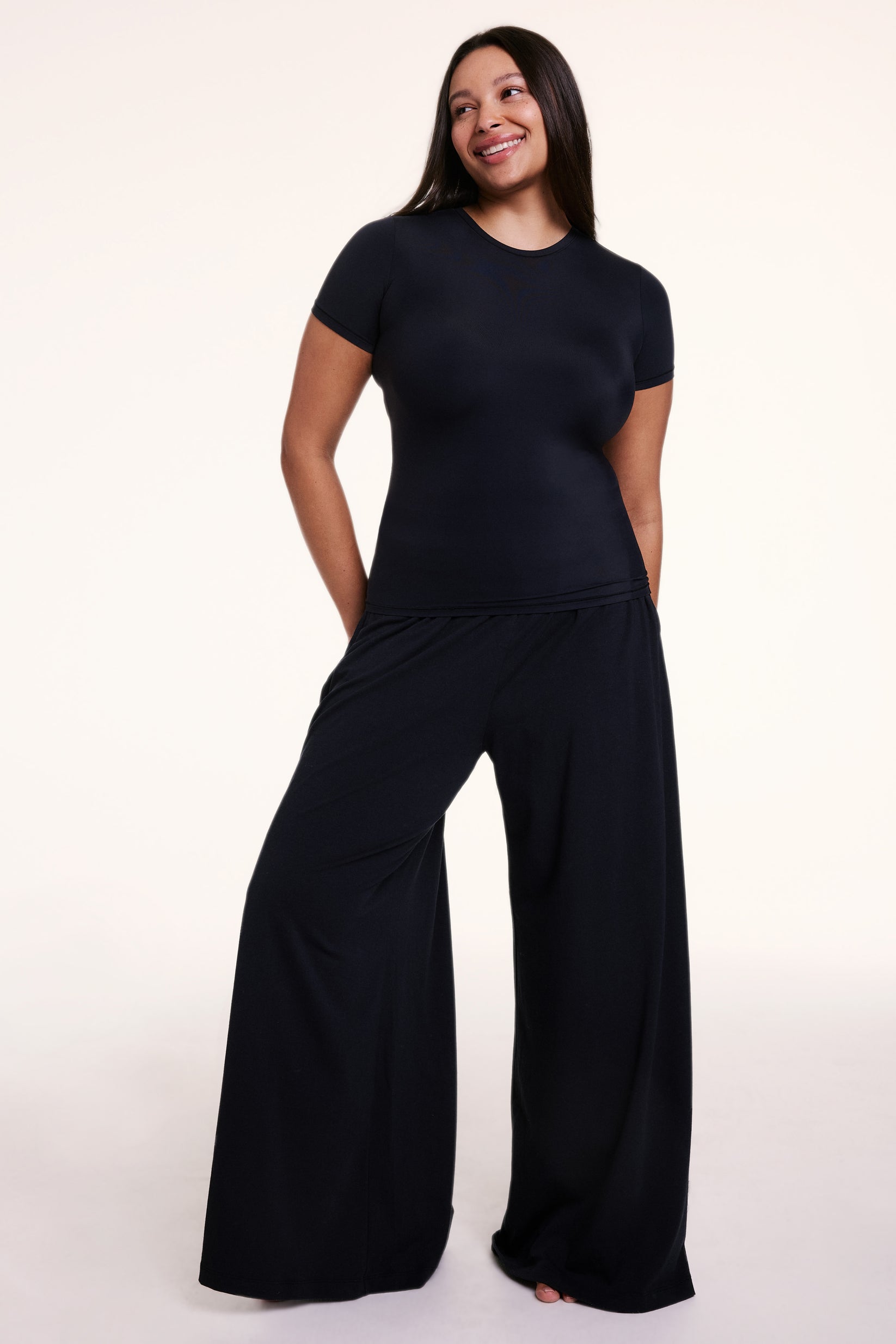 In a studio setting, Alana wears the Bliss Apparel T-Shirt in Black with matching wide-leg pants, featuring a crew neckline, short sleeves, and a smooth, body-hugging fit in ultra-soft microfiber fabric.
