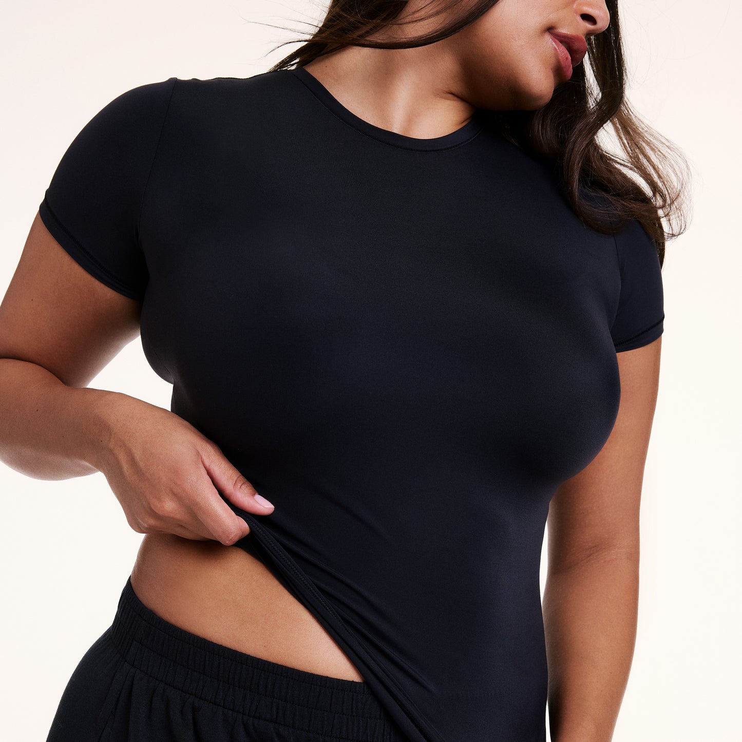 In a close-up studio shot, Alana wears the Bliss Apparel T-Shirt in Black, gently pulling the hem to show the soft, stretchy microfiber fabric and flexible fit.