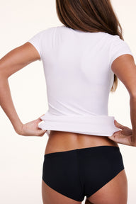 In a studio setting, Linni is shown from the back wearing the Bliss Apparel T-Shirt in White, lifting the hem to highlight the soft, flexible microfiber fabric and close fit.