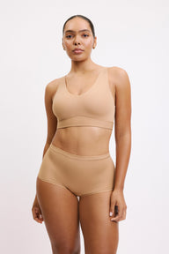 On a white studio backdrop, Amber wears the Bliss Bralette and Bliss Boyshort in Beige, standing facing forward. The bralette features a scoop neckline, wide straps, and a smooth underband, paired with mid-rise boyshorts in soft, lightweight fabric.