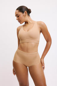 On a white studio backdrop, Amber wears the Bliss Bralette and Bliss Boyshort in Beige, standing at an angle. The bralette features a scoop neckline, wide straps, and a smooth underband, paired with mid-rise boyshorts.