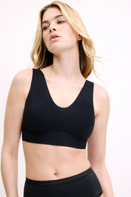 On a white studio backdrop, Megan is shown from a slightly angled front view wearing the Bliss Bralette in Black. The smooth cups and wireless band sit flat under clothing.